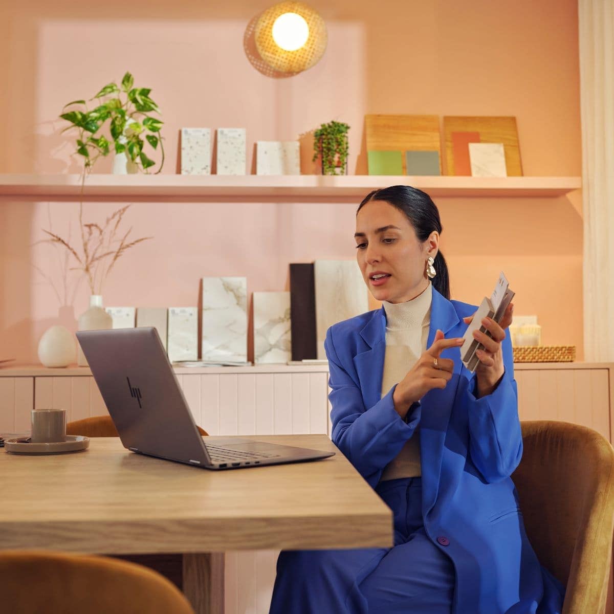 Woman in blue suit working at desk with laptop and phone in stylish pink office. Shelves with plants and decor visible in background